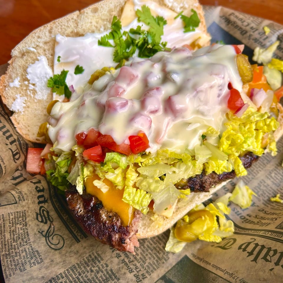 A loaded cheeseburger sub with lettuce, tomatoes, and white sauce, served on newspaper-print paper.