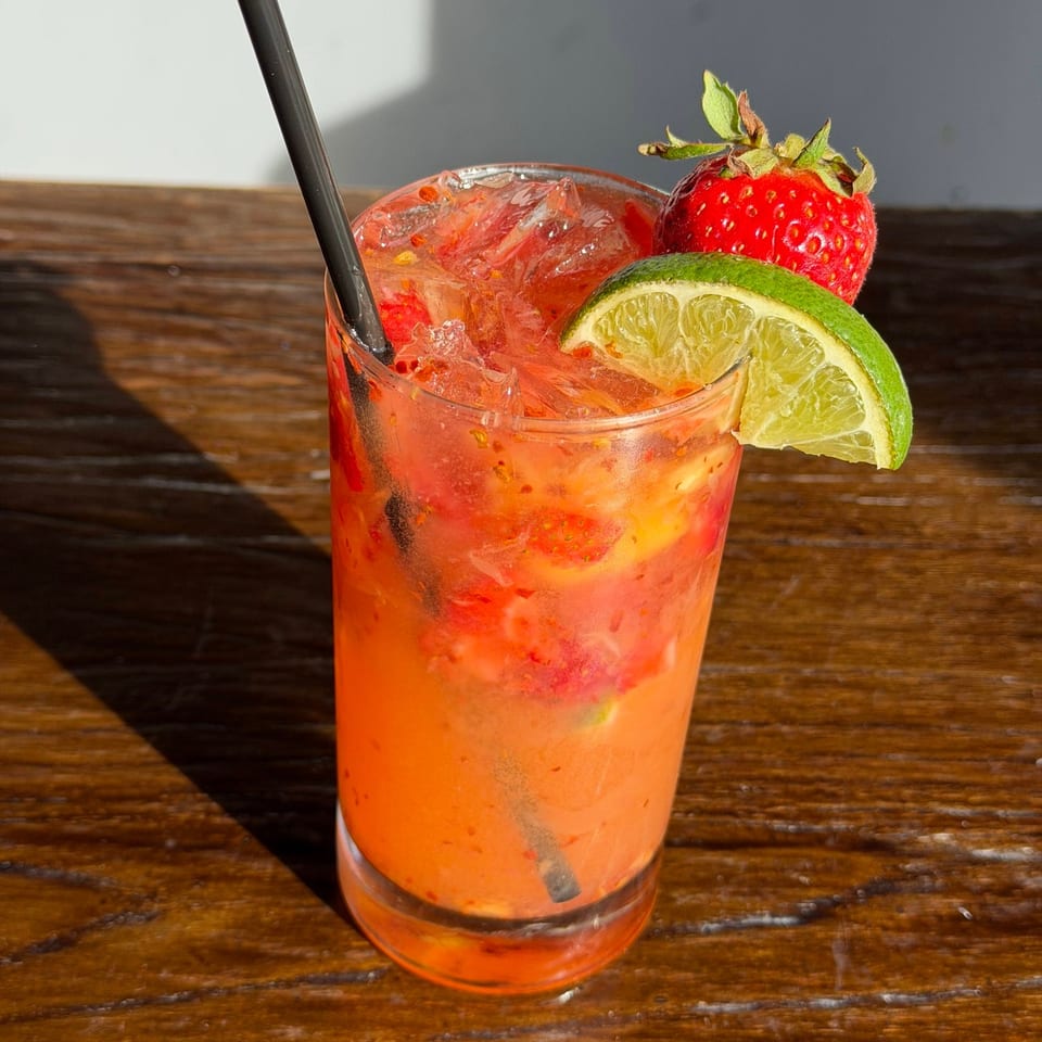 A vibrant strawberry and lime cocktail with a black straw sits on a wooden table.