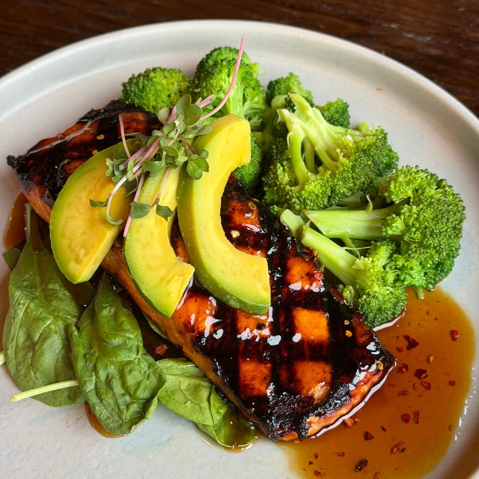 Grilled salmon with avocado slices, broccoli, spinach, and a sweet chili glaze on a white plate.