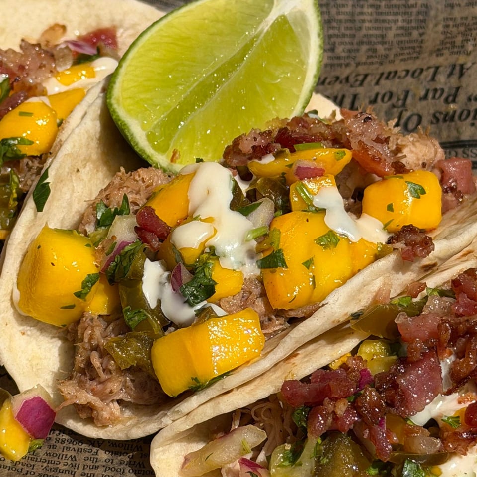 Close-up of three tacos filled with pulled pork, mango salsa, and a lime wedge.