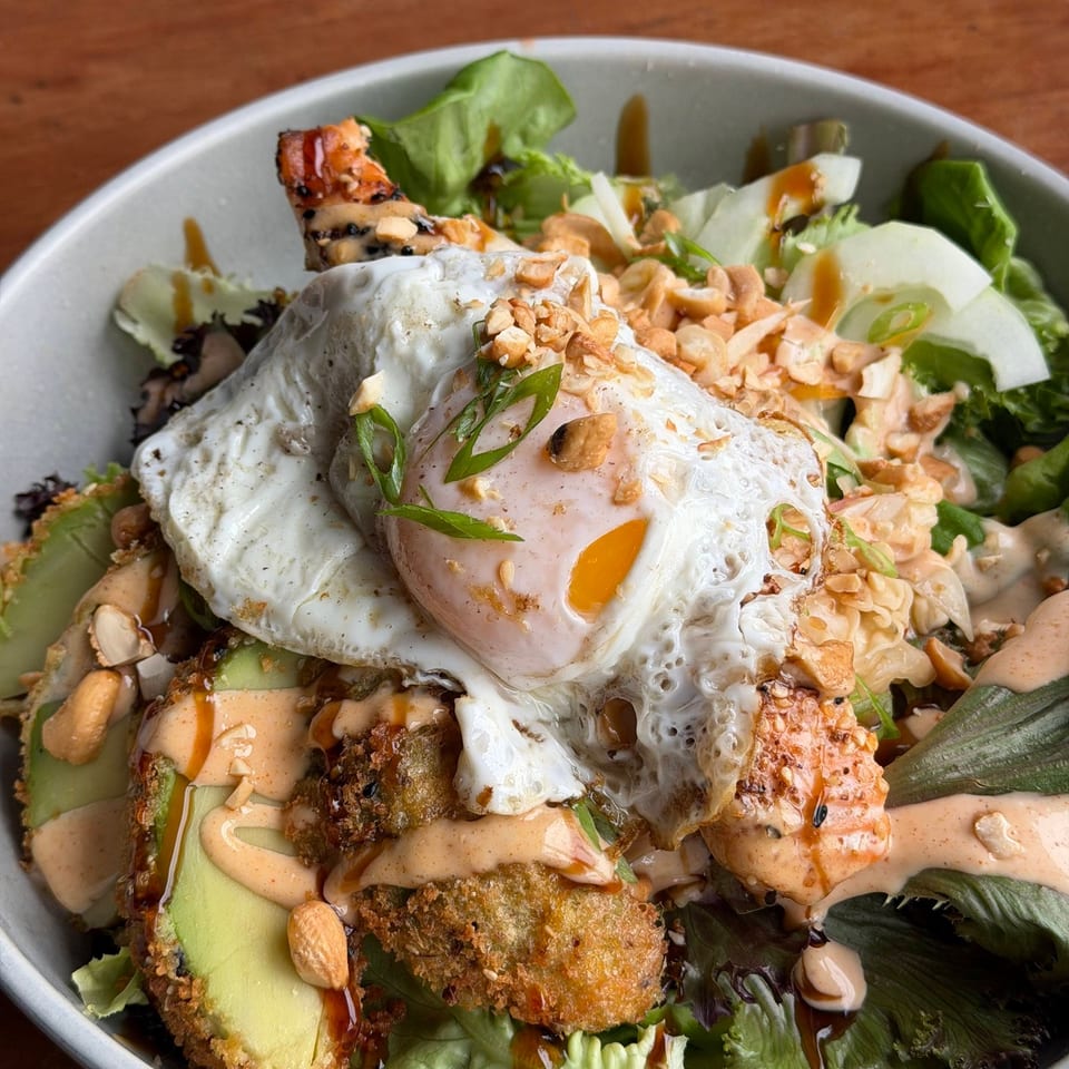 A vibrant salad bowl with fried avocado, shrimp, a fried egg, and a creamy sauce.