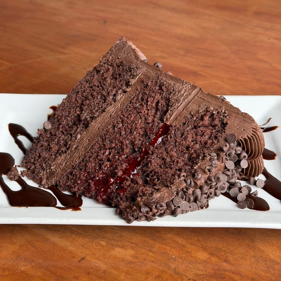 A rich chocolate cake slice with chocolate frosting, raspberry filling, and chocolate chips on a white plate.