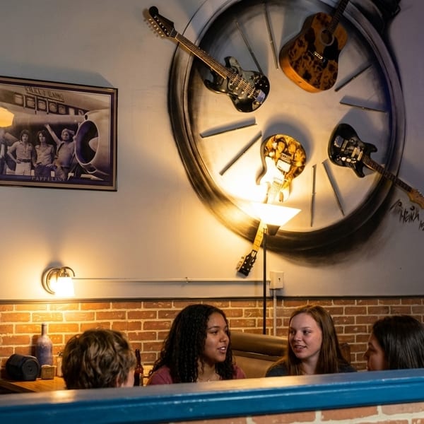 Four people conversing in a restaurant booth with brick walls and guitars decorating the wall.