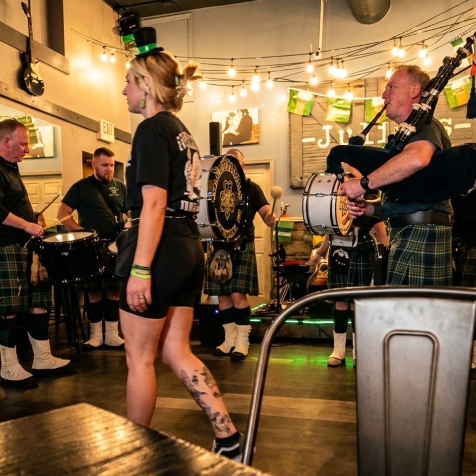 Men in kilts playing bagpipes and drums in a lively pub with string lights and a woman walking by.