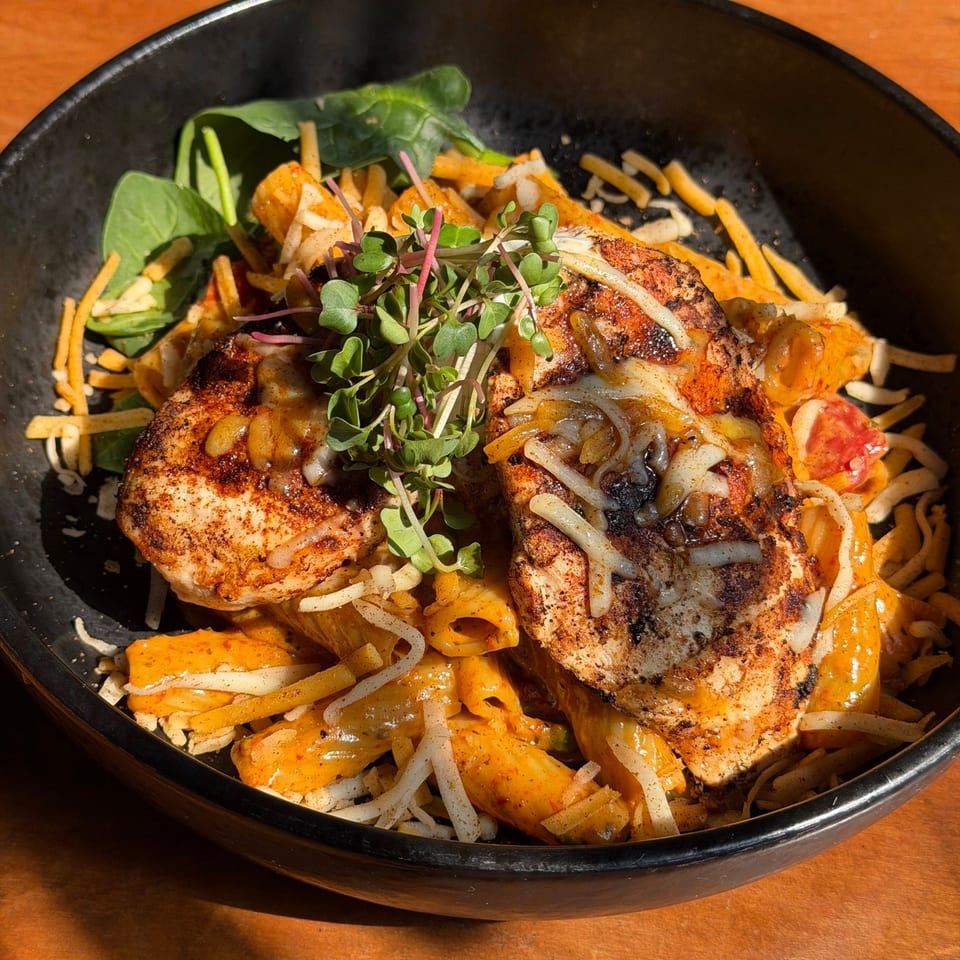 Blackened chicken pasta with melted cheese, spinach, and microgreens in a black bowl.