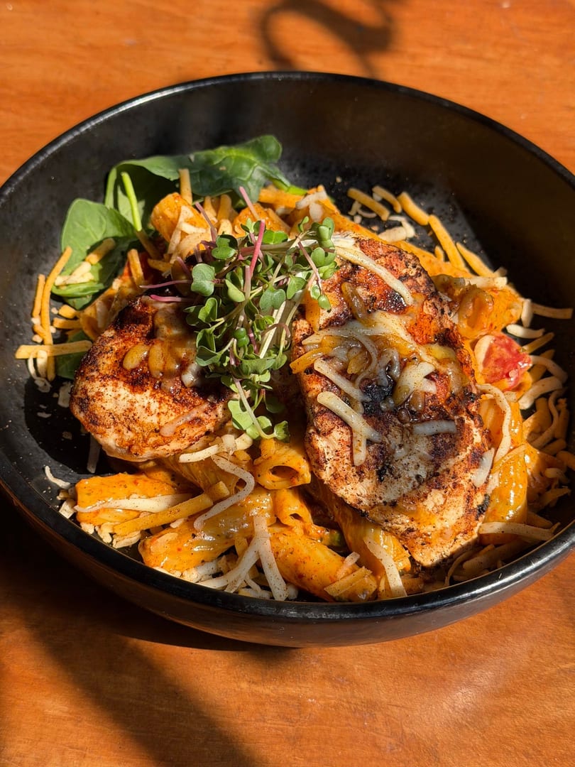 Blackened chicken pasta with melted cheese, spinach, and microgreens in a black bowl.